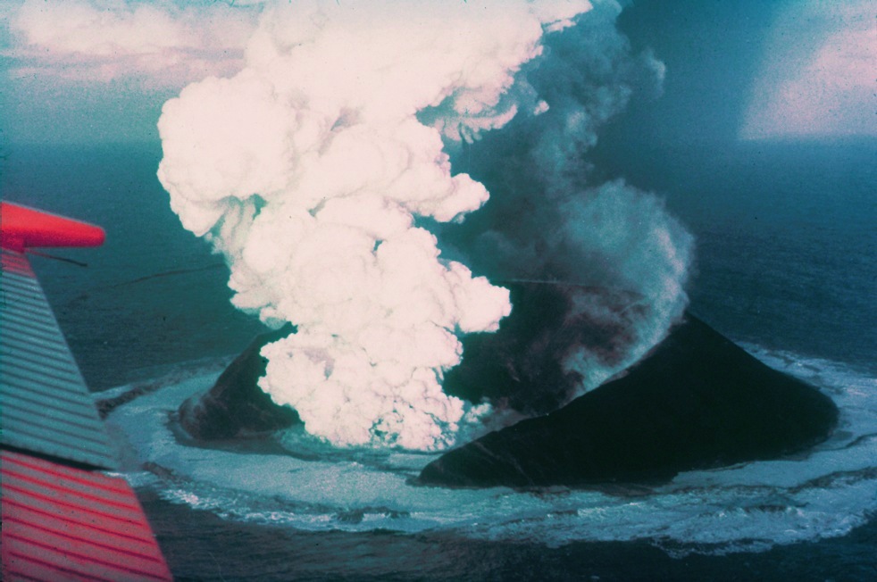 Aerial view of Surtsey erupting on November 30, 1963, fourteen days after its emergence, showing its half-submerged crater from which a plume of volcanic gases and water vapor escapes.